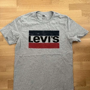 Grå Levi's t-shirt med logga - Snygg grå t-shirt från Levi's med klassisk logga i svart, marinblått och rött framtill. T-shirten är kortärmad och har en rund halsringning. Perfekt för en avslappnad och cool stil.