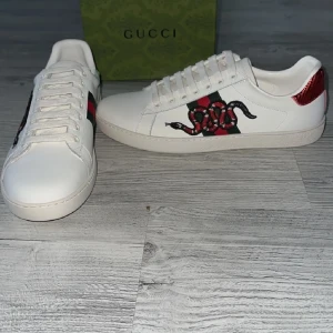 Gucci Ace sneakers med orm - Gucci Ace sneakers i vitt skinn med klassiska gröna och röda ränder på sidan och broderad orm. Baksidan har metallicdetaljer i rött och grönt med Gucci-logga. Snygg och ikonisk modell med rund tå och snörning.