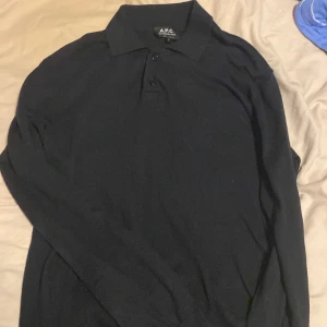 Svart pikétröja från A.P.C. - Klassisk svart långärmad pikétröja från A.P.C. med krage och knappar framtill. Tröjan har en clean och stilren look, perfekt för dig som gillar minimalistisk stil. Tillverkad i mjukt material som känns skönt mot huden.