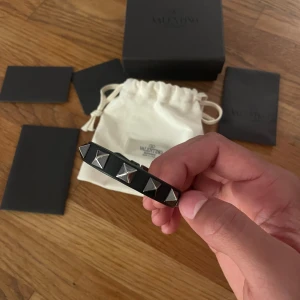 Svart Valentino armband med nitar - Snyggt svart armband från Valentino Garavani med silvriga nitar och klassiskt spänne i metall. Kommer med originalpåse och box. Priset är 1800 men inte hugget i sten kan gå ner i pris om det är snabbt köp