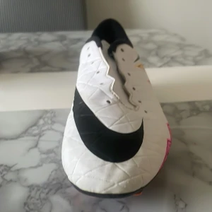 Nike vita fotbollsskor med svart swoosh - Säljer ett par vita Nike fotbollsskor med stor svart swoosh på sidan. Skorna har detaljer i neongult och orange vid hälen och är tillverkade i syntetmaterial. Perfekta för dig som vill sticka ut på planen med en clean och sportig look.