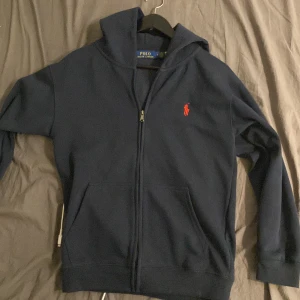 Marinblå hoodie från Polo Ralph Lauren - Säljer en marinblå zip hoodie från Polo Ralph Lauren, använd fåtal gånger, liten defekt som man ser på sista bilden men syns knappt.