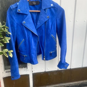 Blå mockajacka från Vero Moda - Snygg blå mockajacka från Vero Moda med klassisk bikerstil. Jackan har silvriga dragkedjor, bred krage och coola detaljer vid axlar och ärmslut. 