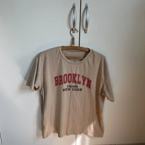 Beige Brooklyn t-shirt med tryck - Snygg beige t-shirt med rött och svart Brooklyn-tryck på bröstet. Skön och mjuk i bomull, enkel att matcha med jeans eller shorts. Storlek L använd fåtal gånger !