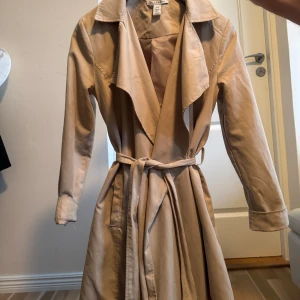 Beige trenchcoat från Na-Kd - Snygg beige trenchcoat från Na-Kd med klassisk krage, långa ärmar och bälte i midjan. Jackan har en avslappnad passform och är perfekt för lager-på-lager. Tillverkad i mjukt material som känns skönt mot huden. Storlek 34-38.