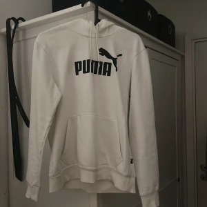 Vit hoodie från Puma med logga - Säljer en stilren vit hoodie från Puma med svart logga och text på bröstet. Tröjan har huva med snörning, magficka och långa ärmar. Perfekt för dig som gillar sportig och avslappnad stil.