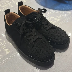Svarta Louboutin sneakers med nitar - Svarta sneakers från Christian Louboutin med coola svarta nitar över tån och klassisk röd sula. Skorna har snörning, rund tå och är gjorda i mocka och skinn. Insidan är beige och mjuk. Perfekta för dig som vill sticka ut med en edgy vibe.