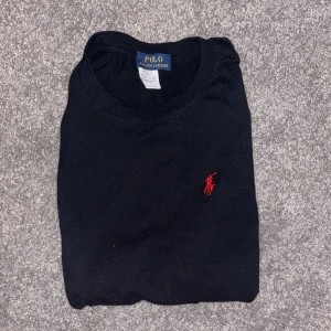 Svart t-shirt från Polo Ralph Lauren - Svart t-shirt från Polo Ralph Lauren med klassisk rund hals och korta ärmar. T-shirten har den ikoniska röda broderade loggan på bröstet och är gjord i storleken M