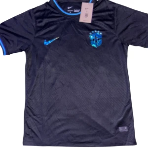 Svart Brasilien Nike fotbollströja - Snygg svart Brasilien fotbollströja från Nike med blå detaljer och holografiskt blått klubbmärke samt Nike-logga på bröstet. Tröjan har korta ärmar och är tillverkad i ett lätt, andningsbart funktionsmaterial. Perfekt för dig som vill sticka ut på planen.