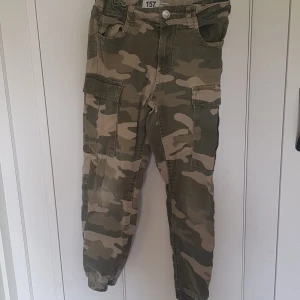 Cargobyxor i camomönster från 157 - Snygga cargobyxor från 157 i klassiskt camomönster i gröna och beige toner. Byxorna har flera praktiska fickor på sidorna och baktill samt resår vid bensluten för en skön passform. Perfekta för en avslappnad och cool stil.