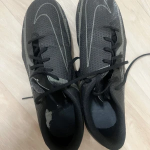 Nike fotbollsskor svart/grå strl 39 - Säljer ett par svarta Nike fotbollsskor med grå detaljer och stora Swoosh-loggor. Skorna har snörning och plös i mesh, samt dobbar för bra grepp på planen. Ovandelen är i syntetmaterial och designen är slimmad och sportig. Perfekta för dig som vill sticka ut på matchen. Endast testade ute en gång