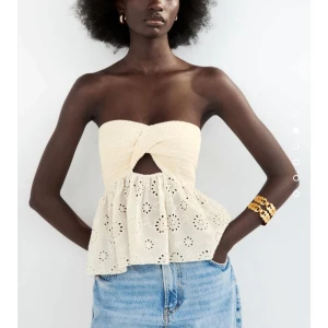 Krämvit bandeau topp från Zara - Superfin bandeau topp i krämvitt från Zara med twistad, ribbstickad överdel och broderad, volangig nederdel i bomull. Toppen är axelbandslös och har ett litet hål framtill för extra detalj. Perfekt till jeans eller kjol för en chill sommarlook.