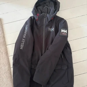 Svart vindjacka från Helly Hansen - Svart vindjacka från Helly Hansen med huva, dragkedja och stora loggor på bröst, ärm och rygg. Jackan har röda detaljer vid huvan och praktiska fickor med tryckknappar.