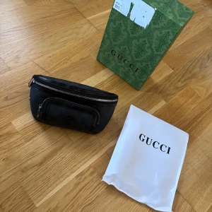 Svart Gucci midjeväska  - Snygg svart magväska från Gucci med klassiskt GG-monogram över hela väskan. Väskan har silverfärgade dragkedjor, justerbart band och ett extra fack framtill. Perfekt för att bära det viktigaste med stil. Kommer med originalpåse och box.