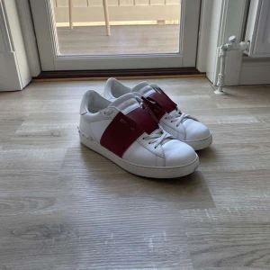 Valentino Open sneakers vit/röd skinn - Snygga Valentino Open sneakers i vitt skinn med bred röd läderdetalj över vristen. Klassisk rund tå, platt sula och diskreta nitar baktill. Skorna har snörning och en stilren design som sticker ut med den röda kontrasten.skick 9/10 priset är ej hugget i sten