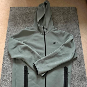 Nike tech khaki grön med luva - Snygg khaki grön tröja från Nike med svart dragkedja och huva. Jackan har två praktiska fickor med dragkedja framtill och diskret Nike-logga på bröstet. Perfekt för dig som gillar sportig och clean stil.