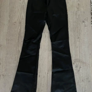 Svarta bootcut byxor från ONLY - Snygga svarta bootcut byxor från ONLY med glansig finish. Byxorna har klassisk femficksdesign, bälteshällor och stängs med knapp och dragkedja. Perfekta för dig som gillar en trendig och stilren look.