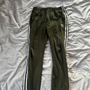 Olivgröna Adidas mjukisbyxor med ränder - Snygga olivgröna mjukisbyxor från Adidas med klassiska vita ränder längs sidorna och broderad logga framtill. 