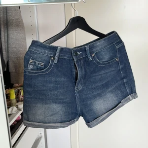 Jeansshorts med broderi och paljetter - Mörkblå jeansshorts med uppvikta benslut och snygga broderade bakfickor dekorerade med paljetter. Klassisk femficksmodell med silverfärgade nitar och detaljer. Materialet är 85% bomull och 15% polyester, vilket gör dem både mjuka och slitstarka. FEJK MISS ME. DMa för mer info🩷