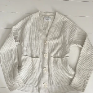 Vit ribbad kofta från Zara - Vit ribbstickad kofta från Zara med stora knappar framtill och två praktiska fickor. Koftan har lång ärm och en klassisk v-ringning. Perfekt för lager-på-lager och enkel att matcha med det mesta. Har blivit lite lite nopprig på ena ärmen men ska fixa det innan den säljs