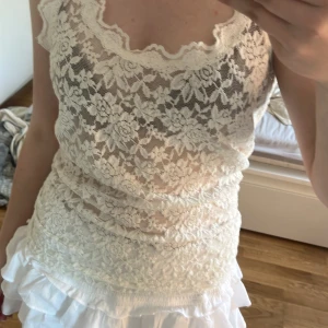 Vit spetsig linnetopp med blommönster - Säljer en söt vit linnetopp i spets med blommigt mönster och vågig kant vid urringningen. Toppen är ärmlös och har en romantisk känsla, perfekt för varma dagar eller lager på lager-stil.
