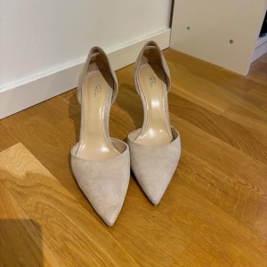 Beige mockapumps med spetsig tå - Snygga pumps från Rizzo i beige mocka med spetsig tå och hög klack. Skorna har en stilren och elegant design med öppen insida och klassisk siluett. Perfekta för dig som vill ha en clean och chic look.