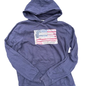 Blå hoodie Ralph Lauren Denim & Supply, USA flag - Mörkblå hoodie från Ralph Lauren Denim & Supply med stor amerikansk flagga-patch framtill. Tröjan har huva, magficka och långa ärmar. Tillverkad i mjuk bomull, perfekt för chill dagar. Snygg vintagekänsla med slitna detaljer på flaggan. Inget fel med tröjan, inga hål, inga fläckar. Hoodien är äkta. Storleken är L men passar även utmärkt till M också