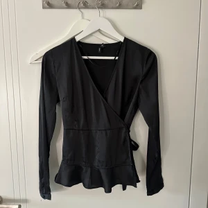 Svart omlottopp med volangkant - Snygg svart omlottopp från vero Moda med långa ärmar och v-ringning. Toppen har knyt i sidan och en volangkant nedtill som ger en extra touch❤️