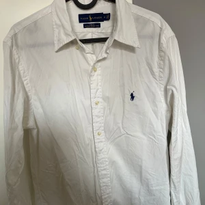 Vit skjorta från Ralph Lauren - Klassisk vit skjorta från Ralph Lauren med blå broderad logga på bröstet. Skjortan har lång ärm, knappar framtill och är tillverkad i tunn bomull. Perfekt för en clean och stilren look.