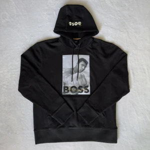 Svart hoodie BOSS x G.O.A.T. - Svart hoodie från BOSS med tryck på bröstet och texten 'BOSS' samt 'G.O.A.T.' på ryggen. Huva med snörning och tryckt logga. Klassisk passform och mjukt material, perfekt för en cool streetstyle-look.