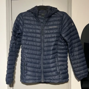 Mörkblå dunjacka Peak Performance - Snygg mörkblå dunjacka från Peak Performance med huva och dragkedja framtill. Jackan är quiltad med horisontella sömmar och har två fickor med dragkedja. Perfekt för kyliga dagar och enkel att matcha med olika outfits. Jackan är i storlek 170.