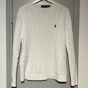Vit kabelstickad tröja Polo Ralph Lauren - Vit kabelstickad tröja från Polo Ralph Lauren med rund halsringning och broderad marinblå logga på bröstet. Tröjan har långa ärmar och ribbade muddar för en klassisk look. Perfekt för lager-på-lager under kyliga dagar. Den är i storlek M och passar normalt till storleken.