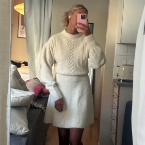 Stickad kortklänning från Adoore - Supermysig stickad klänning från Adoore med kabelstickat mönster framtill, rund hals och långa puffärmar. Klänningen är kort, har markerad midja och är gjord i mjukt stickat material. Obs är den i vit!