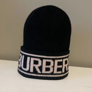Svart Burberry mössa med logga - Svart ribbstickad mössa från Burberry med bred uppvikt kant där Burberrys logga syns i beige. Klassisk och stilren design som ger en lyxig känsla. Perfekt för dig som vill ha en snygg accessoar med tydlig branding.