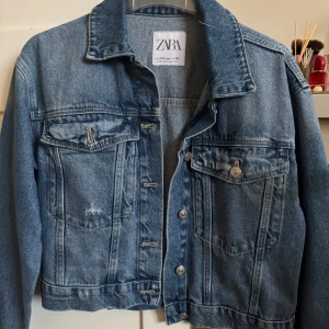 Blå jeansjacka från Zara, stl 152 - Klassisk blå jeansjacka från Zara med snygga silverskimrande knappar och två bröstfickor med lock. Jackan har en rak passform, krage och långa ärmar. Jackan är i 152 men passar även xxs
