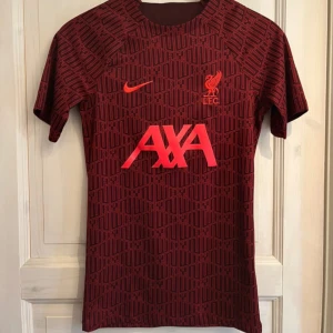 Liverpool  - Liverpool FC tränings-tshirt från Nike i vinröd färg med svart geometriskt mönster. AXA-logga och klubbmärke i rött framtill. Tillverkad i Dri-FIT-material som andas, perfekt för fotbollsträning. Kortärmad och slim fit-modell. Köpt på unisport för 999kr