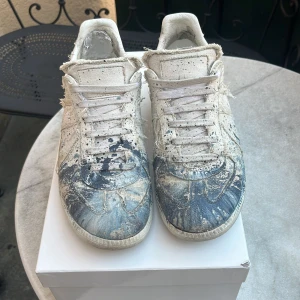 Maison Margiela sneakers med färgstänk - Unika sneakers från Maison Margiela i vitt canvas med blåfärgstänk och slitna detaljer. Skorna passar nån som har en unik och kreativ stil hör av om frågar eller annat har några defekter 