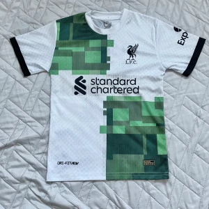 Liverpool Salah bortatröja 22/23 Nike - Liverpool FC bortatröja säsongen 22/23 med Mo Salah och nummer 11 på ryggen. Tröjan är vit med gröna och mörkgröna grafiska blockdetaljer, svarta tryck och svarta muddar. Tillverkad i lätt Dri-FIT-material som andas. Perfekt för dig som älskar fotboll, Liverpool eller Salah. Storlek är 152 men passar folk från ca 150 - 155cm.