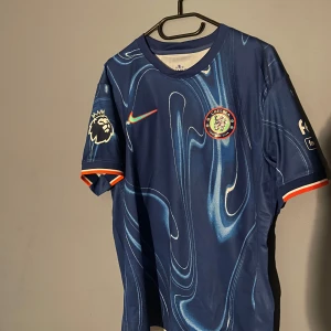 Chelsea Madueke 11  - Chelsea FC matchtröja med Madueke 11-tryck. Djupblå bas med unikt mönster, klubbmärke på bröstet och Premier League-logga på ärmen. Orange och vita detaljer vid ärmslut. Tillverkad i lätt och ventilerande material, perfekt för fotbollsfans.