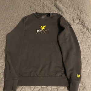 Svart sweatshirt från Lyle & Scott - Svart sweatshirt från Lyle & Scott med klassisk gul logga på bröstet och ärmen. Tröjan har rund hals, ribbade muddar och är tillverkad i mjuk bomull. Perfekt för en clean och stilren look med tydlig streetkänsla.
