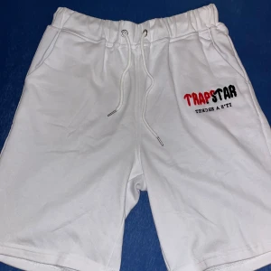 Vita Trapstar shorts - Snygga vita shorts från Trapstar med broderad logga i rött och svart framtill. De har elastisk midja med snörning och två sidofickor. Perfekta för chill dagar eller när du vill ha en clean streetwear-look.