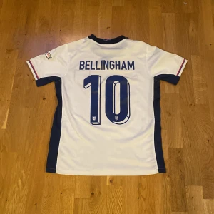 England fotbollströja Bellingham 10 - England herrlandslagets vita fotbollströja med blå detaljer, nummer 10 och Bellingham på ryggen. Broderat landslagsmärke på bröstet och EM-märke på ärmen. Klassisk V-ringning och korta ärmar. Perfekt för fans. Pris kan diskuteras.