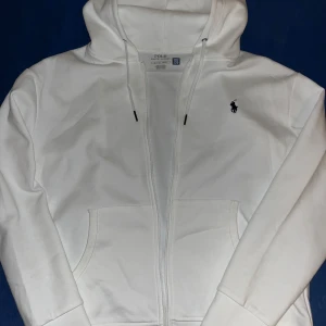 Vit kofta från Polo Ralph Lauren - Vit kofta från Polo Ralph Lauren med dragkedja. Tillverkad i mjuk bomull, perfekt för sommaren.