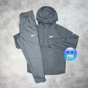 Nike miller tracksuit  - Nike träningsset i grått med jacka och byxa. Jackan har huva, dragkedja och Nike-logga på bröstet. Byxorna har resår i midjan och logga på låret. Perfekt för löpning eller chill. Lätt och smidig polyester som andas.