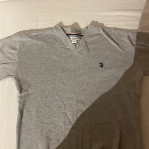 Grå v-ringad tröja från Polo Assn - Snygg grå v-ringad tröja från U.S. Polo Assn. med broderad logga på bröstet. Tröjan har en klassisk passform och är tillverkad i mjuk bomull, perfekt för lager-på-lager. Stilren och enkel att matcha med det mesta.