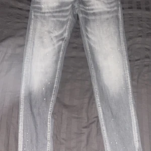 Grå Dsquared2 Jeans - Snygga grå jeansbyxor från Dsquared2 med coolt blekt tvättad look och vita färgstänk över hela byxan. Klassisk femficksmodell med svarta nitar och röd Dsquared2-tag vid gylfen. Raka ben och normal passform, perfekt för en streetinspirerad stil. 10/10 skick. Längd 100cm Slim fit.