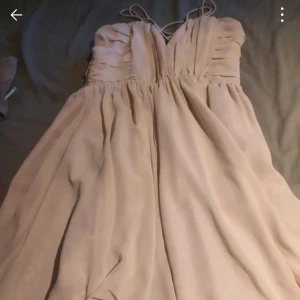 Champagnefärgad långklänning med snörning - Elegant långklänning i champagnefärg med v-ringad omlottliv och tunna axelband med snörning i ryggen. Klänningen har ett luftigt, svepande fall och draperad överdel som ger en romantisk känsla. Perfekt för bal eller student.