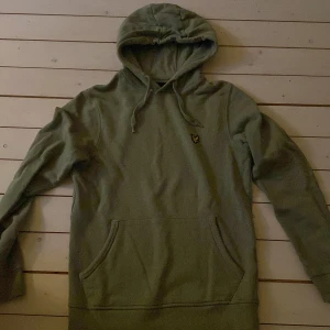 Olivgrön hoodie från Lyle & Scott - Olivgrön hoodie från Lyle & Scott med klassisk gul logga på bröstet. Tröjan har huva med snörning, känguruficka och långa ärmar. Tillverkad i mjuk bomull, perfekt för chill dagar eller när du vill ha en enkel men snygg look.