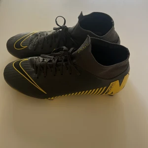 Nike Mercurial fotbollsskor svart/gul - Snygga Nike Mercurial fotbollsskor i svart med gula detaljer och ränder. Skorna har hög strumpliknande krage i flyknit-material, snörning och plös med FLYKNIT-tryck. Perfekta för dig som vill sticka ut på planen med stil och grym passform.