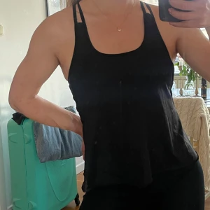 Svart linne med inbyggd sportbh - Säljer en svart linne med smala dubbla axelband som korsas i ryggen och inbyggd sportbh. Toppen har en lös passform och är tillverkad i ett mjukt, stretchigt material som passar perfekt till träning